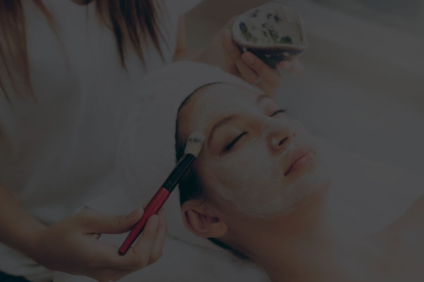 Person lying down with eyes closed, a towel wrapped around their head, receiving a facial treatment.